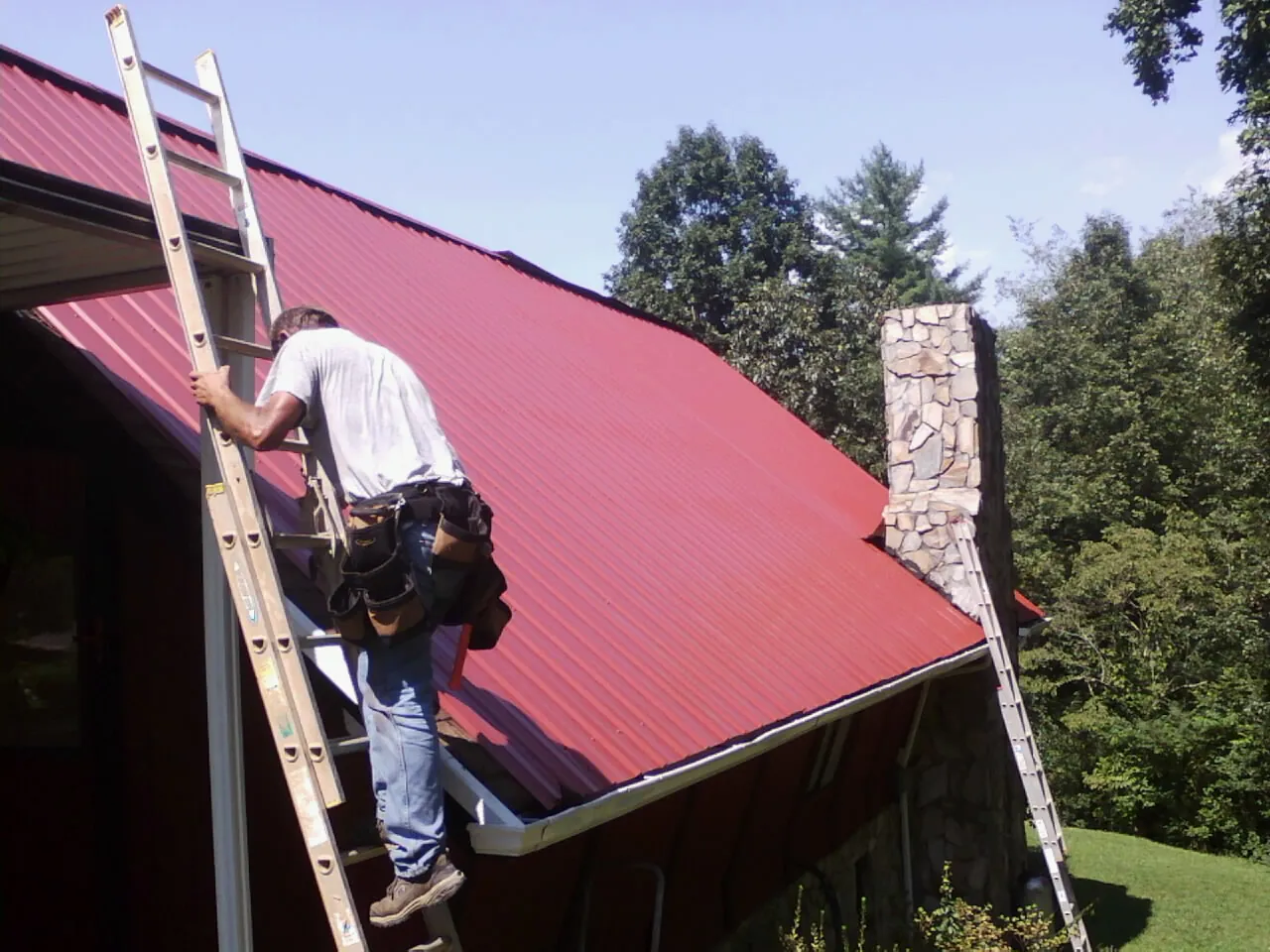Roofer on a ladder beside a red metal roof and stone chimney — Soffit & Fascia Repair by Roofing Brothers in New Canton